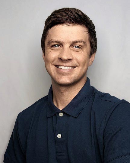 The image shows a smiling man with short hair, wearing a blue shirt, posing for a portrait with a neutral background.