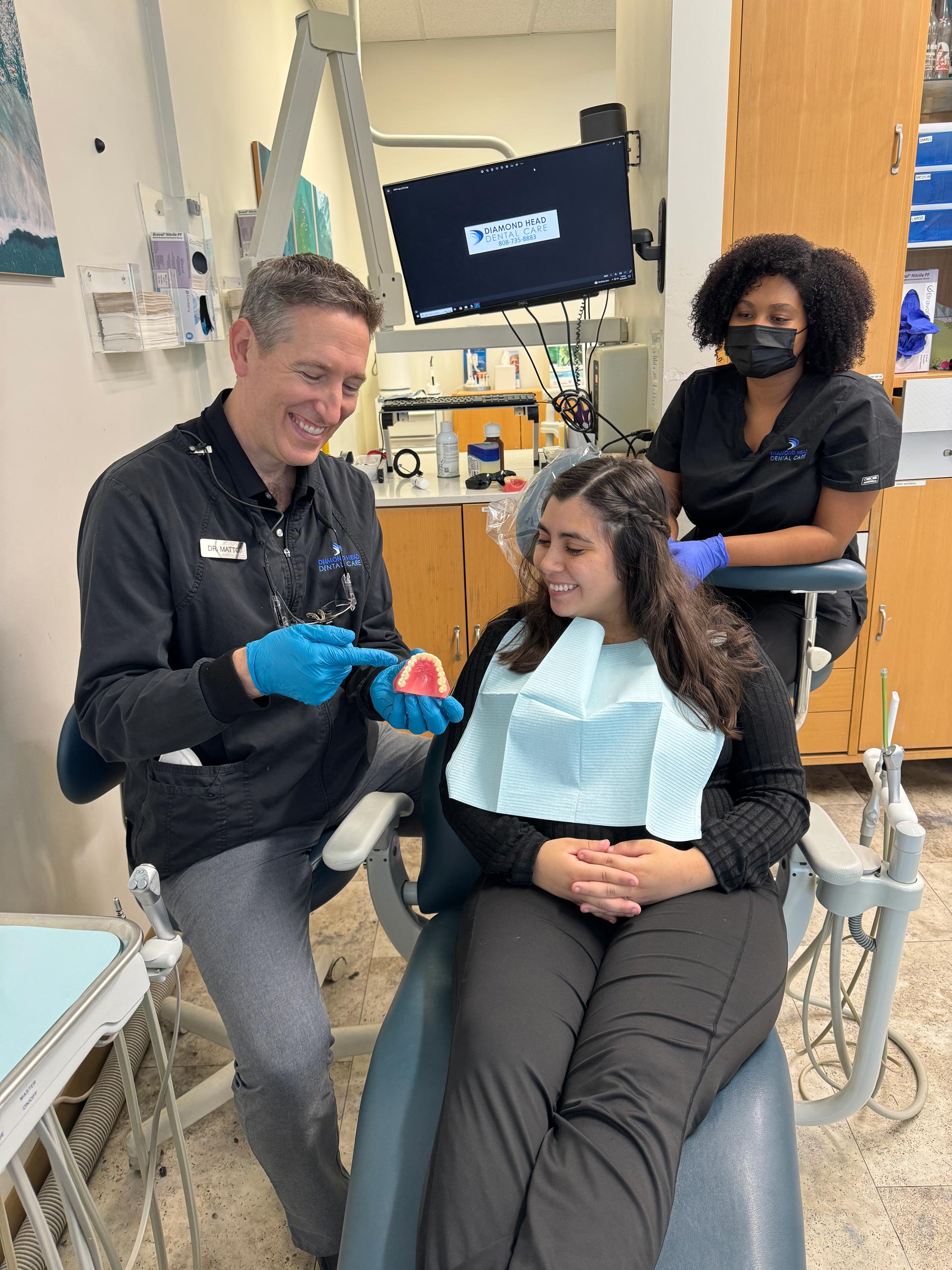 A dental professional examines a patient's teeth while two other individuals observe, with medical equipment visible in the background.