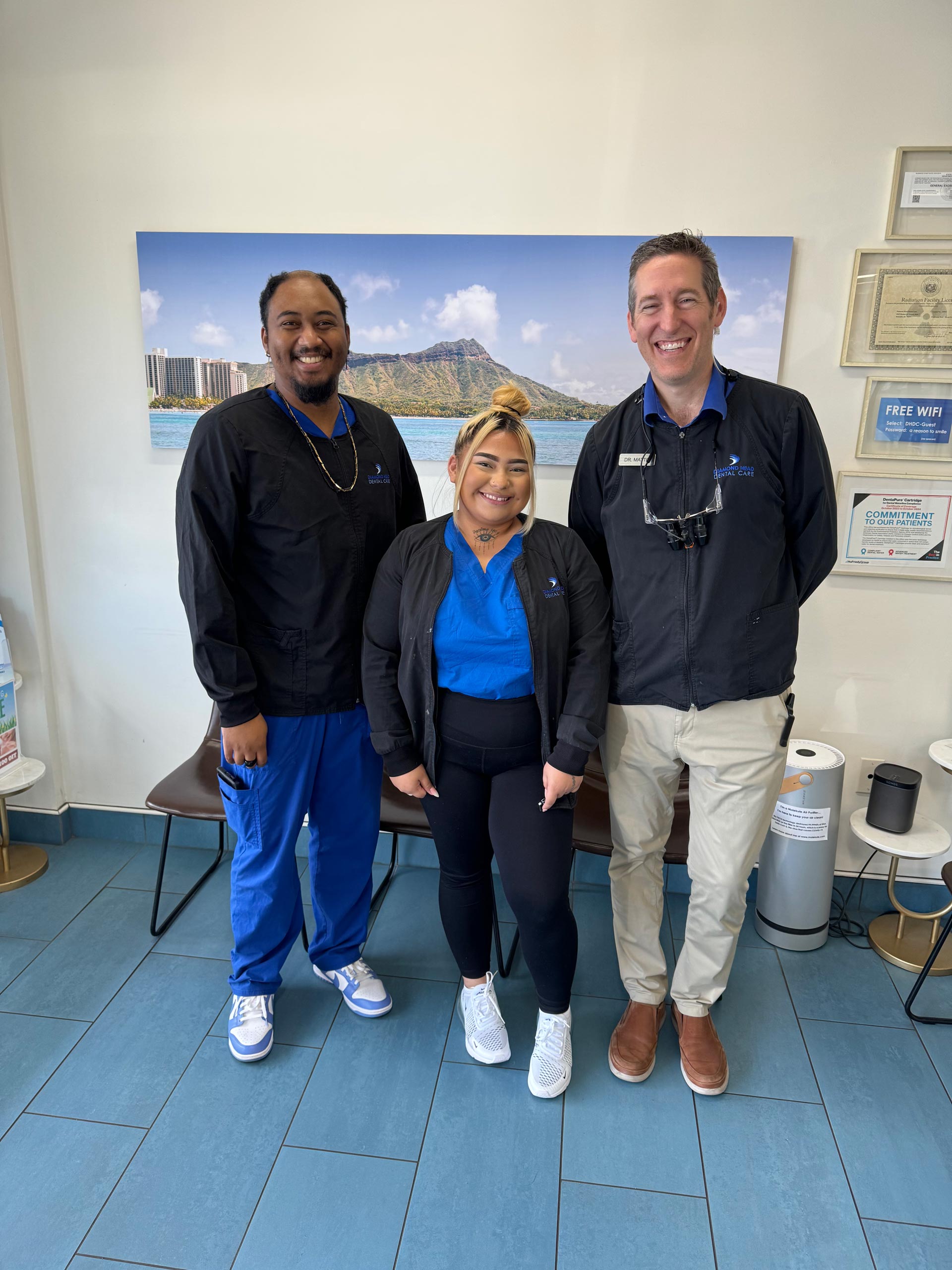 Three individuals posing in front of a wall-mounted picture with a scenic background, standing in an office setting.