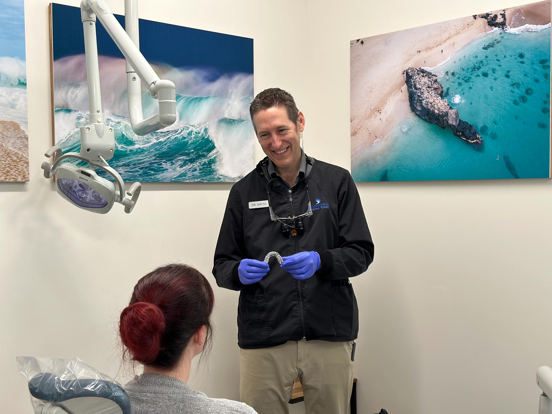 A dentist in a dental office, standing next to a patient, smiling at the camera.