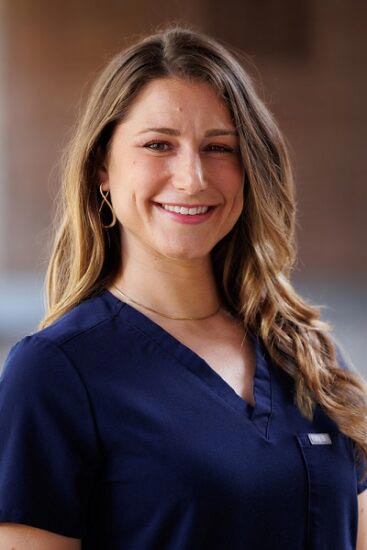 The image shows a woman wearing a blue scrub top, smiling at the camera, with her hair styled in loose waves.