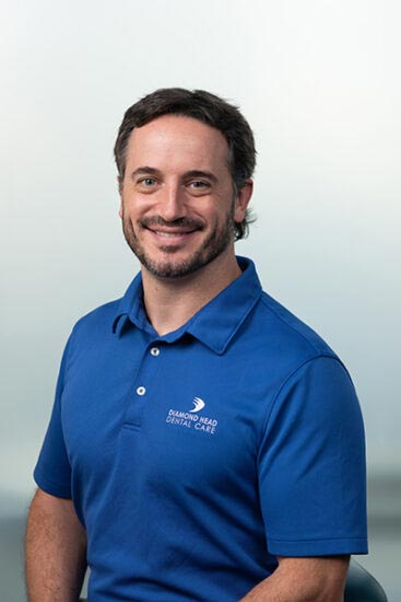 The image shows a man wearing a blue polo shirt standing against a light background. He has short hair, a beard, and is smiling at the camera.