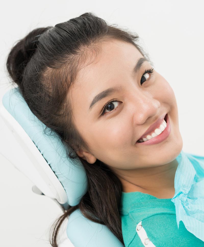 A young woman with a bright smile poses with a dental chair, showcasing her teeth and the dental equipment.