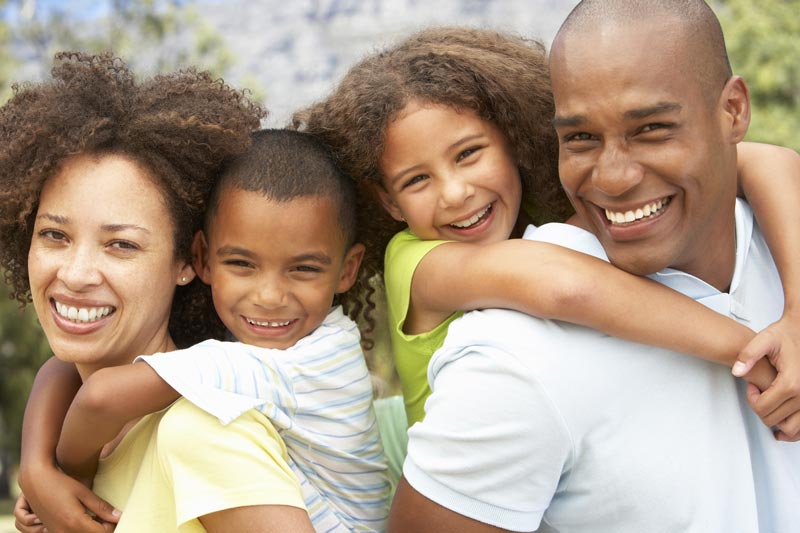 The image depicts a family of four, consisting of an adult male, an adult female, and two children, all smiling and embracing each other outdoors during daylight.