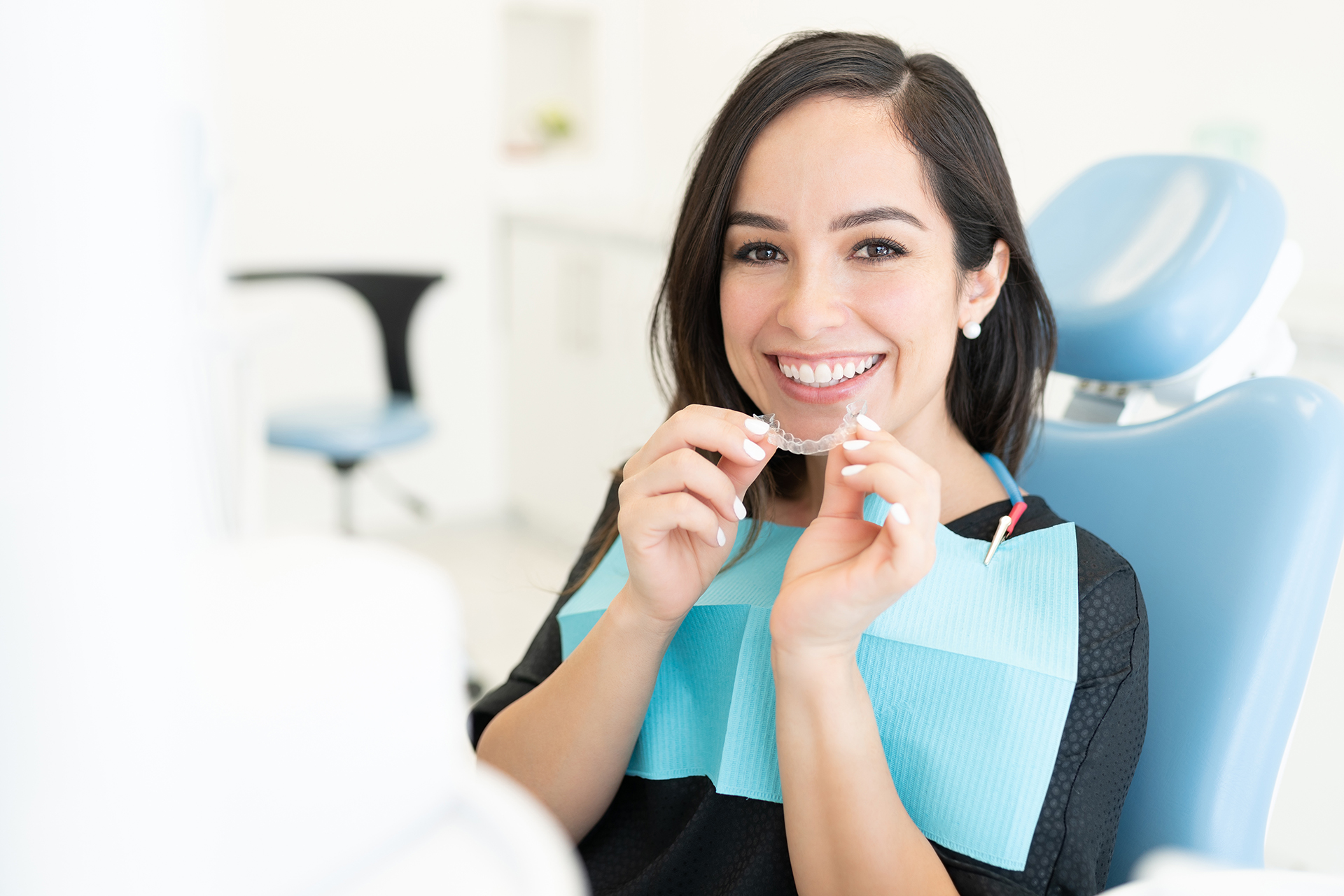 The image shows a smiling woman sitting at a dental chair with a tray of dental tools in front of her, wearing a black shirt, and holding a toothbrush in her hand.