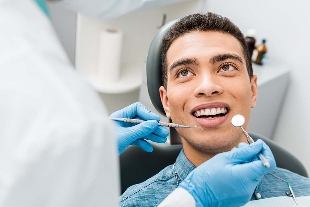 The image depicts a man sitting in a dental chair with his mouth open, receiving treatment from a dentist who is working on him, while wearing protective gloves and holding tools.