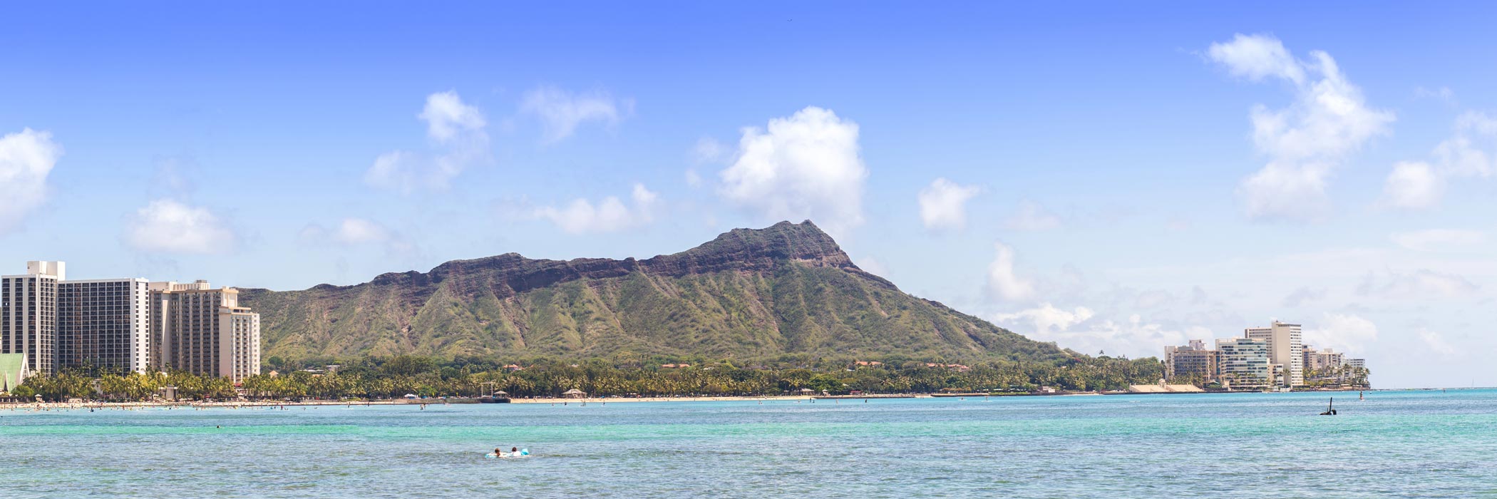 The image displays a panoramic view of a tropical island with clear blue skies and a cityscape in the distance, with palm trees visible along the beachfront.