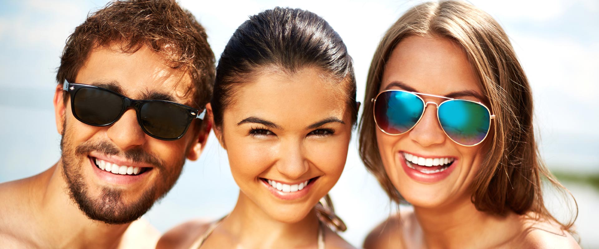 A group of four people posing together with smiles, wearing sunglasses and swimwear, standing on a sandy beach.