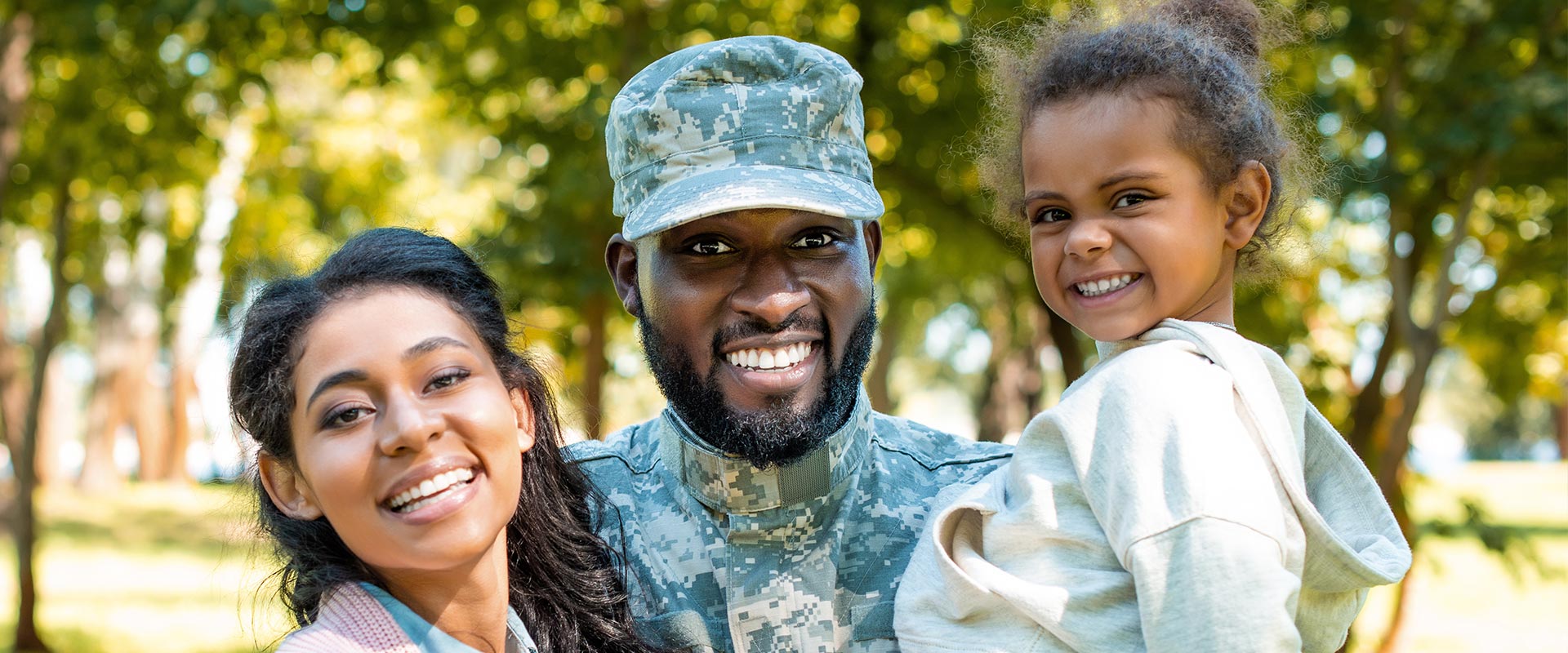 The image features a family of three posing with a man in military uniform, all smiling and standing together outdoors during daylight.