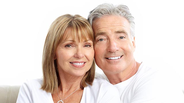 The image shows two adults, an older man and woman, standing close together and smiling at the camera. They are posing for a portrait with a neutral background.