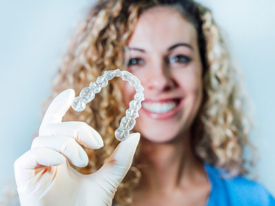A woman holding up a clear plastic dental retainer with her left hand, smiling at the camera.