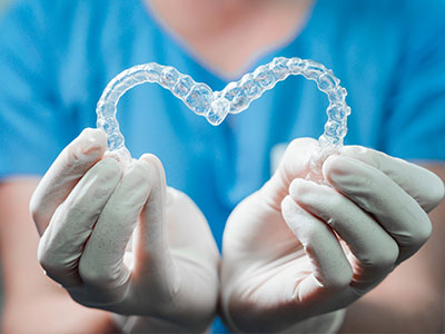 The image shows a hand holding a pair of clear braces with a heart shape on them against a blue background.