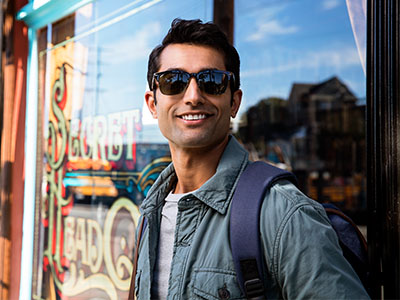 A young man with sunglasses stands confidently outside a storefront, wearing a backpack and a light-colored jacket.
