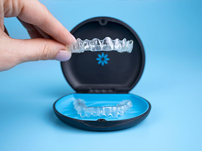 The image shows a hand holding an open package containing a clear retainer with a blue lid, set against a blue background.