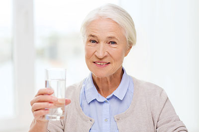 The image shows an elderly woman holding up a glass of water with both hands, smiling at the camera, and wearing a light blue top.