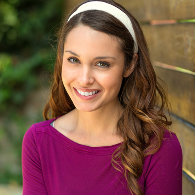 The image shows a woman with long brown hair smiling at the camera while wearing a purple top and a white headband.