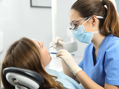 A dental hygienist working on a woman s teeth while she is seated in a dentist s chair, with both of them wearing personal protective equipment.