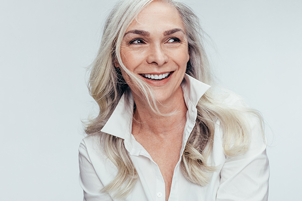 The image shows a woman with short blonde hair smiling at the camera, wearing a white shirt with rolled-up sleeves and her right hand on her chin.