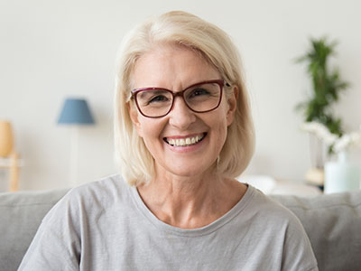 The image shows a woman with blonde hair wearing glasses and a light-colored top, smiling at the camera. She has short hair and appears to be indoors, possibly in a living room setting, as suggested by the presence of a couch and a lamp in the background.