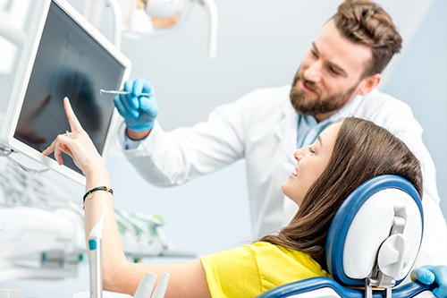 A man and woman in a medical setting, with the man standing behind a patient seated in a chair with an open mouth, examining teeth  they are surrounded by dental equipment and technology.