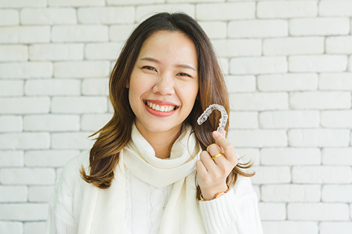 A woman with a smile holds up a hand with a peace sign gesture against a brick wall background.