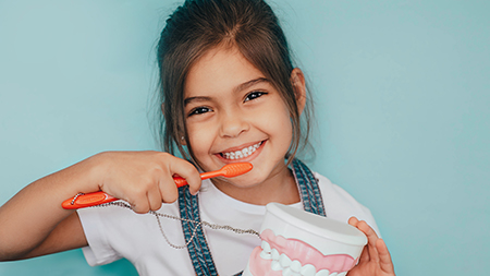 The image shows a young girl with a toothbrush in her mouth, smiling at the camera, holding a cupcake with a bite taken out of it.