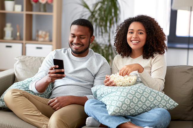 This is a color photograph showing a man and woman sitting on a couch with a relaxed posture, both holding smartphones, and they are smiling at each other while watching something on one of their devices.