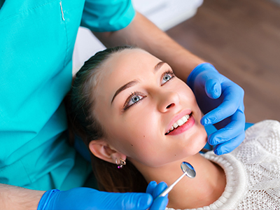 The image shows a person sitting in a dental chair with a dental hygienist performing a cleaning procedure, wearing blue gloves and holding a mirror to inspect the patient's mouth.