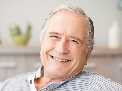 An older man with gray hair, smiling, sitting comfortably in a home setting.
