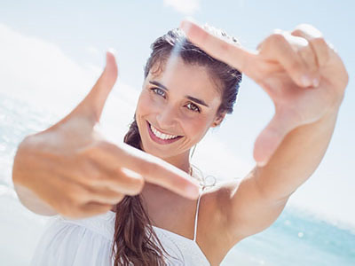 The image shows a woman taking a selfie with her hand held up to her face, making a peace sign gesture. She has long hair, is wearing a white top, and stands against a bright background that suggests an outdoor setting, possibly by the sea.