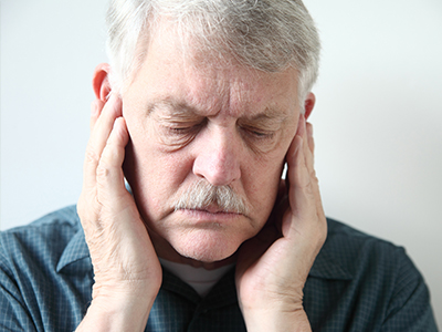 The image shows a man with his hands placed over his ears, appearing to be listening intently or experiencing discomfort, with a neutral background.